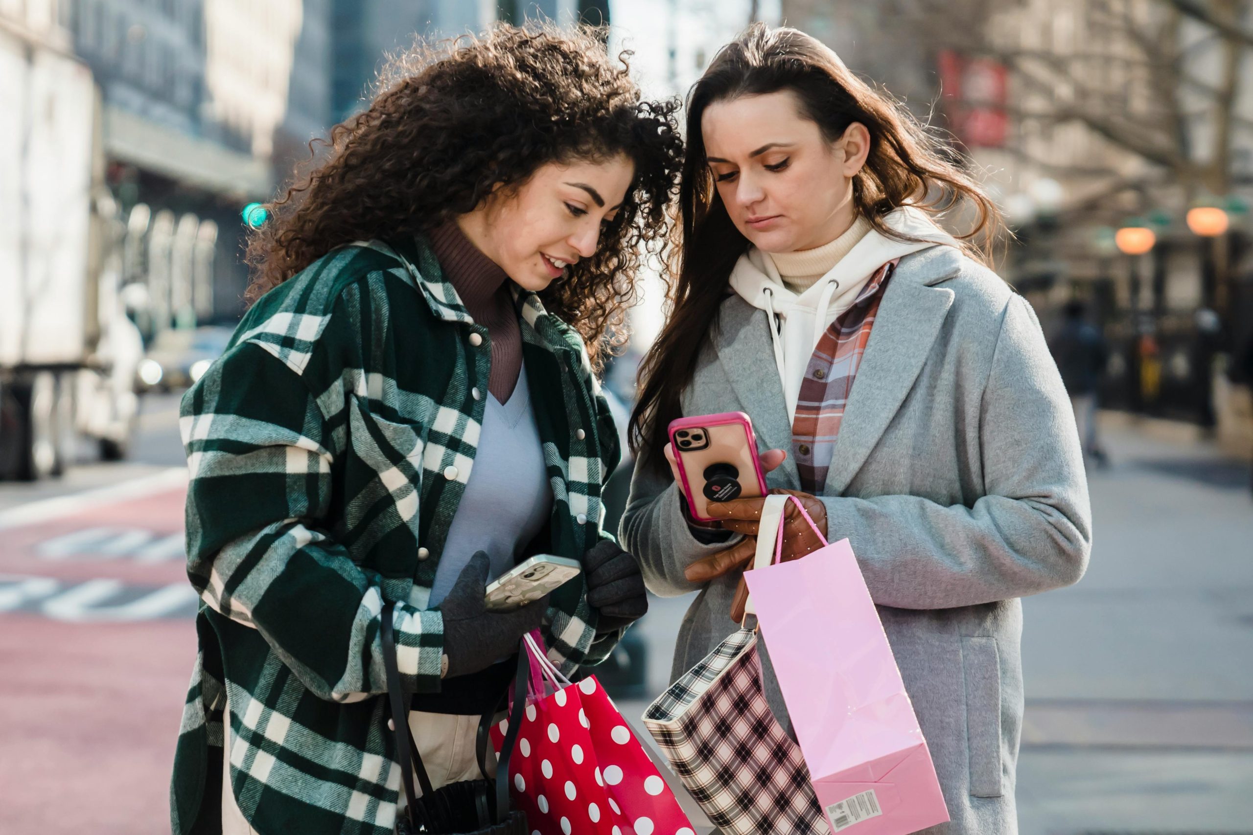 Calm women friends in trendy clothes standing with shopping bags on city street and browsing smartphone in sunny day
