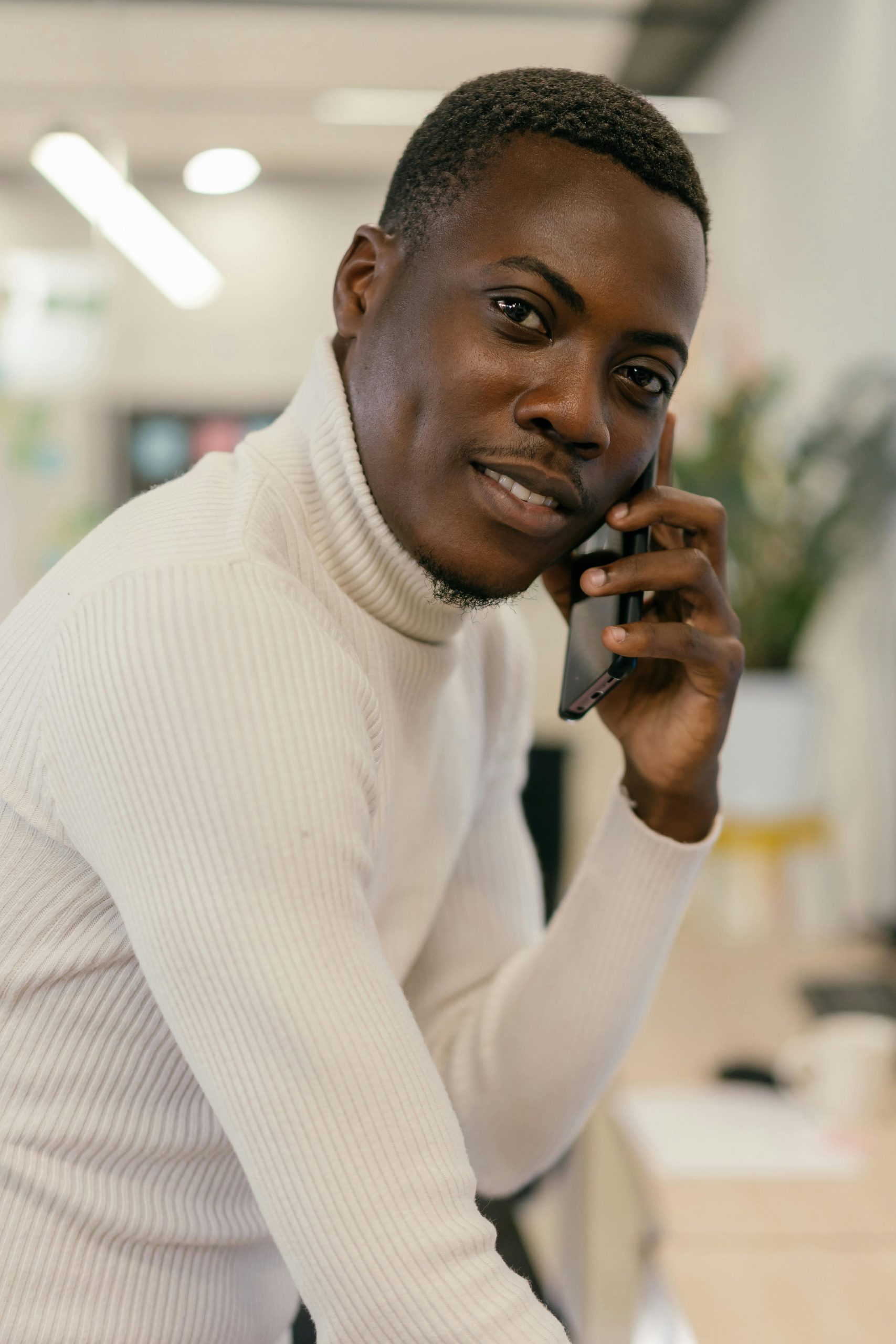 Smiling man in a white sweater making a phone call in a contemporary office setting.